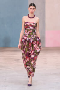 Model walking a runway in a strapless floral purple dress with a chunky purple necklace and matching purple heels.