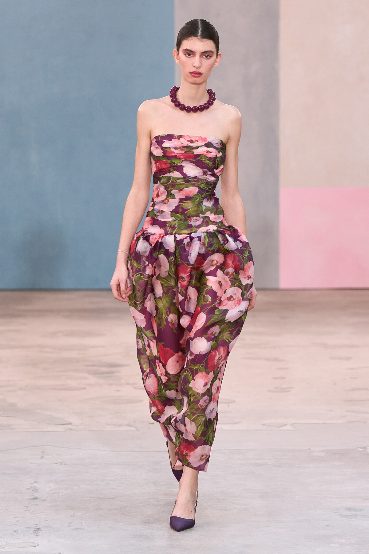 Model walking a runway in a strapless floral purple dress with a chunky purple necklace and matching purple heels.