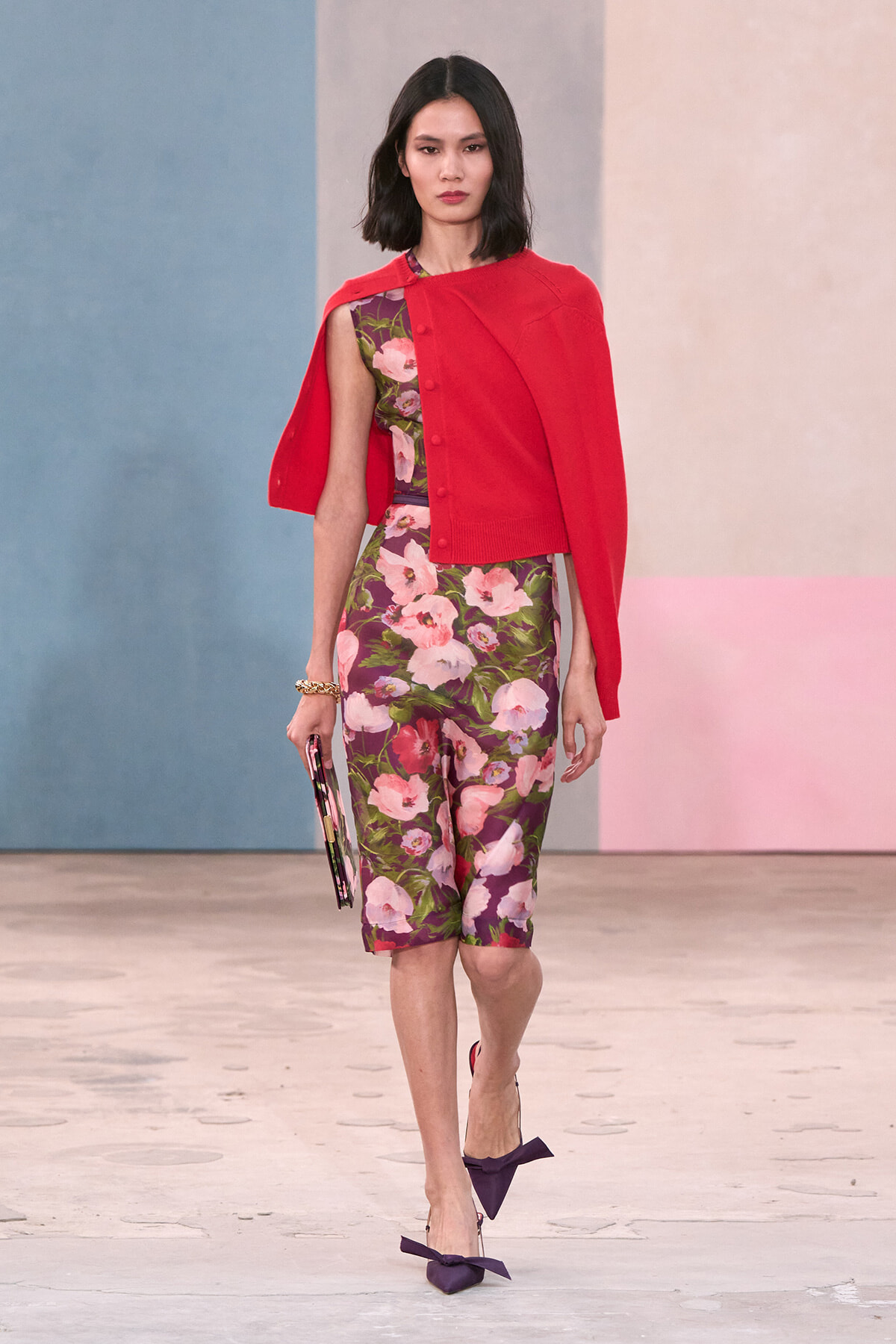 Model on a runway wearing a red cape over a pink-floral dress, carrying a clutch and wearing purple bow-tied heels.