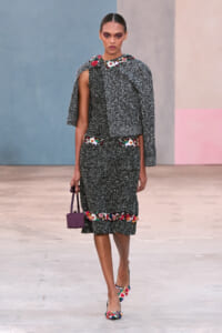 Runway model in a textured black tweed dress with floral embroidery and a cape sleeve, carrying a small purple handbag at her side.