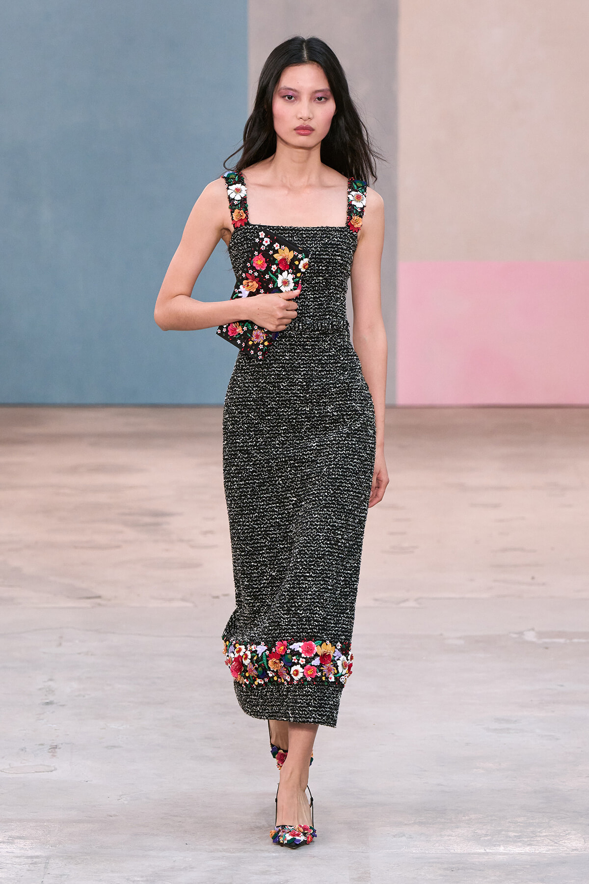 Model walking in a black textured midi dress with colorful floral embroidery on straps and hem, carrying a matching floral clutch on a fashion runway.