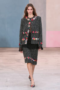 Model on a runway wearing a black tweed suit with colorful floral embroidery along edges and cuffs, paired with a matching skirt and floral heels.