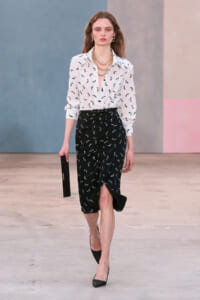 Model walking a runway in a white bird-print blouse and black wrapped midi skirt with a front slit, accessorized with gold chains, earrings, and a black clutch.