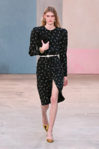 Model on a runway wearing a black midi dress with a white bird print, white belt, and a wrapped skirt with a front slit, holding a black clutch with gold heels.