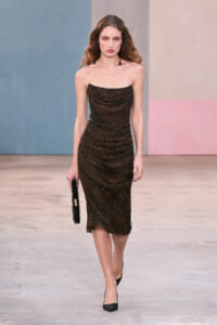 Model walking the runway in a strapless brown leopard-print midi dress, holding a black clutch and wearing black pointed heels.