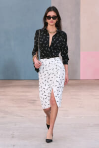 Runway look: black bird-print blouse tucked into a white printed skirt with a front slit, paired with black heels and gold jewelry.