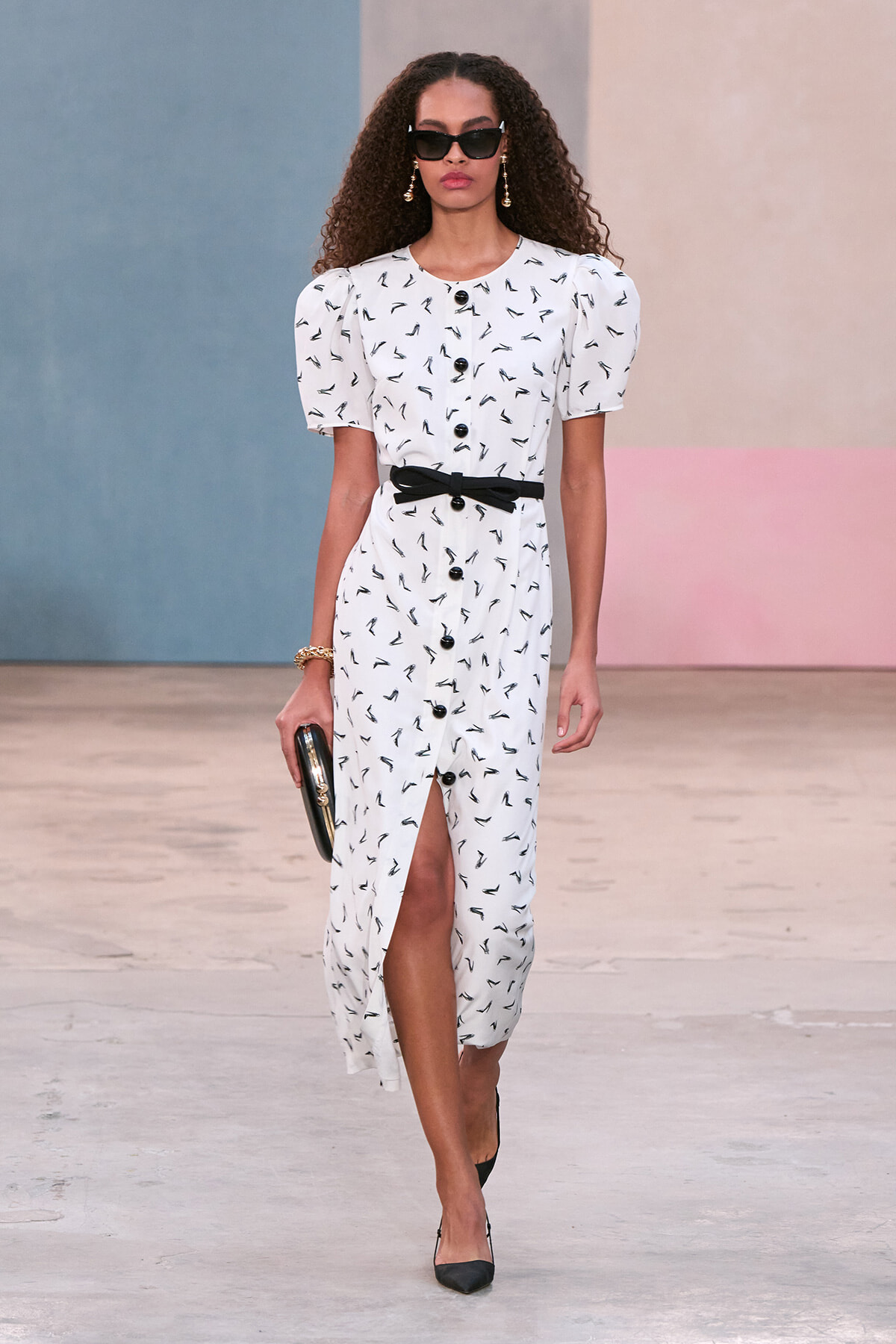 Model walking the runway in a white printed button-down dress with puff sleeves, black belt, and black heels, carrying a clutch and wearing sunglasses.
