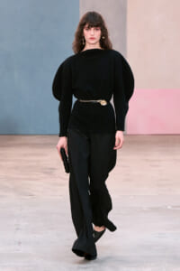 Model on a runway wearing an all-black ensemble with sculpted shoulders, a gold belt, and wide-leg pants, carrying a black clutch and gold earrings.