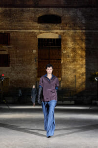 Runway model walks a fashion show on an industrial brick-walled stage, wearing a purple satin top, blue wide-leg pants, and long black gloves.