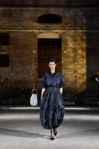 Model walking on a runway in a black, voluminous satin dress in an industrial brick-walled venue; spotlighted center stage with another model in a white skirt in the background.