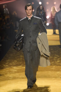 Male model walking a fashion runway in a charcoal grey suit, carrying a beige overcoat and a dark leather bag.