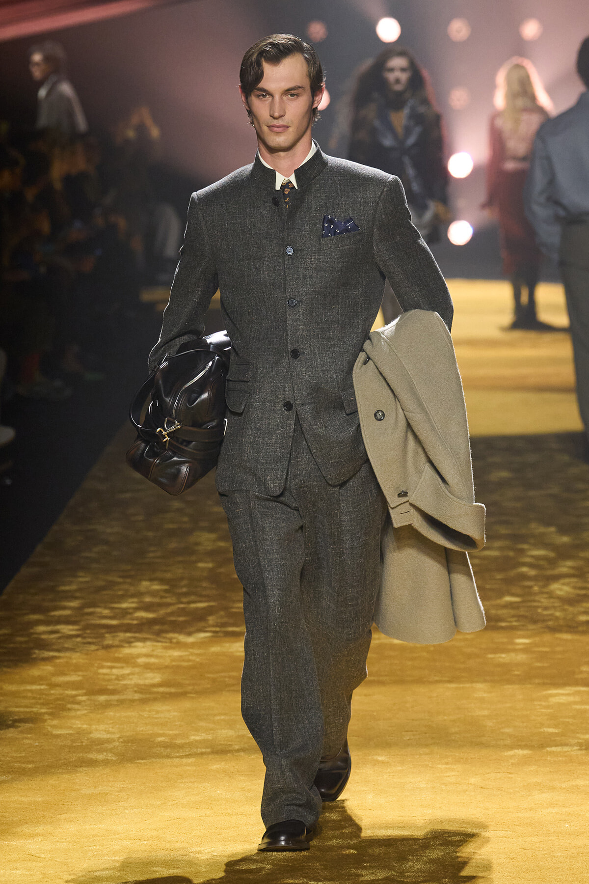Male model walking a fashion runway in a charcoal grey suit, carrying a beige overcoat and a dark leather bag.