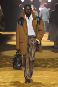 Male model walks a fashion runway wearing a camel coat with black leather accents, striped shirt, brown trousers, and a black leather duffel bag on his arm.