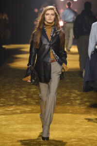 Female model walking a fashion runway in a black leather blazer over a camel turtleneck, with a blue patterned scarf and gray tailored trousers, carrying a dark bag.