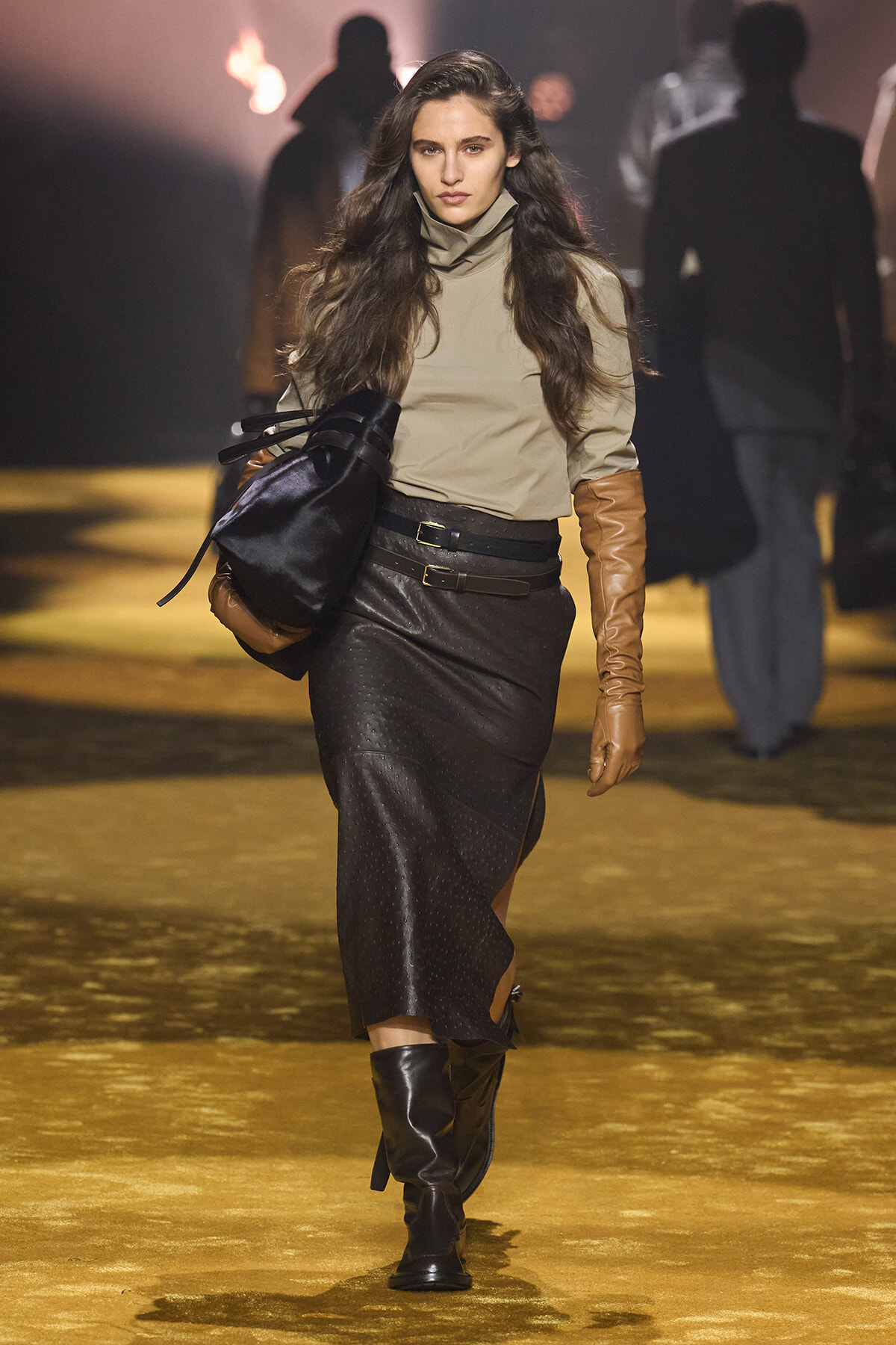 Model walks a runway in a beige turtleneck, black textured midi skirt, brown leather gloves, knee-high boots, and carries a black bag.