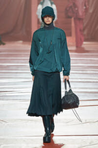 Model walking a runway in a teal sporty-hooded windbreaker, matching pleated skirt, teal cap, black boots, and a black drawstring handbag.