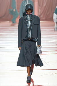 Runway model wearing a dark oversized suit with a hooded jacket, cap, and a pleated midi skirt, holding a small silver handbag.