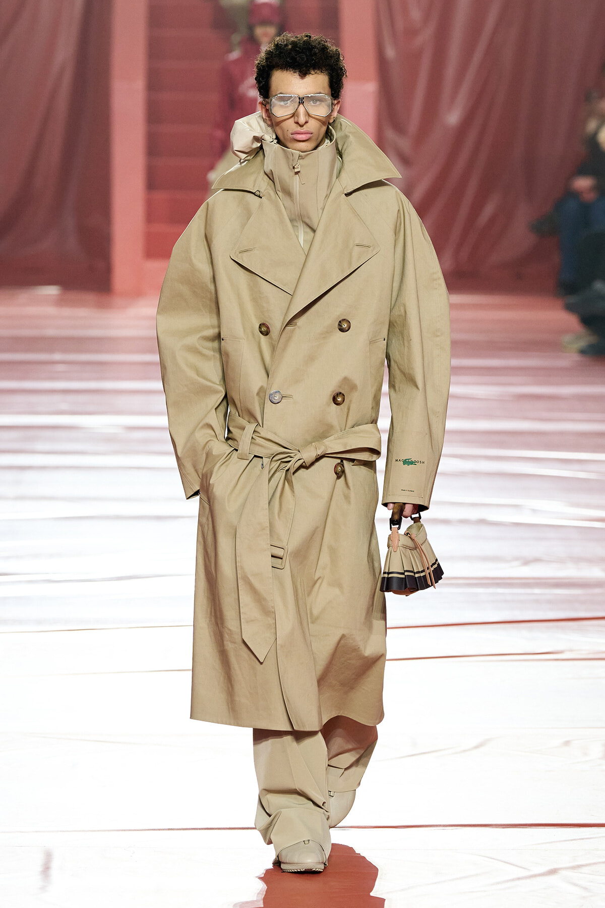 Male model on a fashion runway wearing an oversized beige trench coat with a waist belt, large sunglasses, and a small striped handbag.