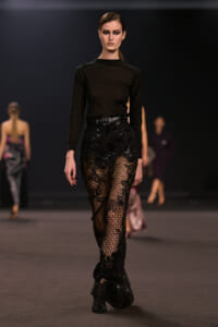 Model walks a runway in a black long-sleeve top and sheer black lace trousers with sequins, belted at the waist.