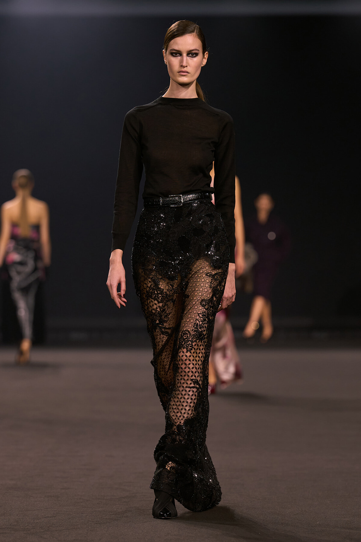 Model walks a runway in a black long-sleeve top and sheer black lace trousers with sequins, belted at the waist.
