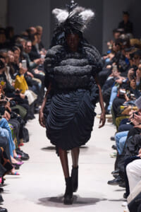 Model walks the runway in a dramatic black sculptural dress with a voluminous quilted bodice and feathered headpiece.
