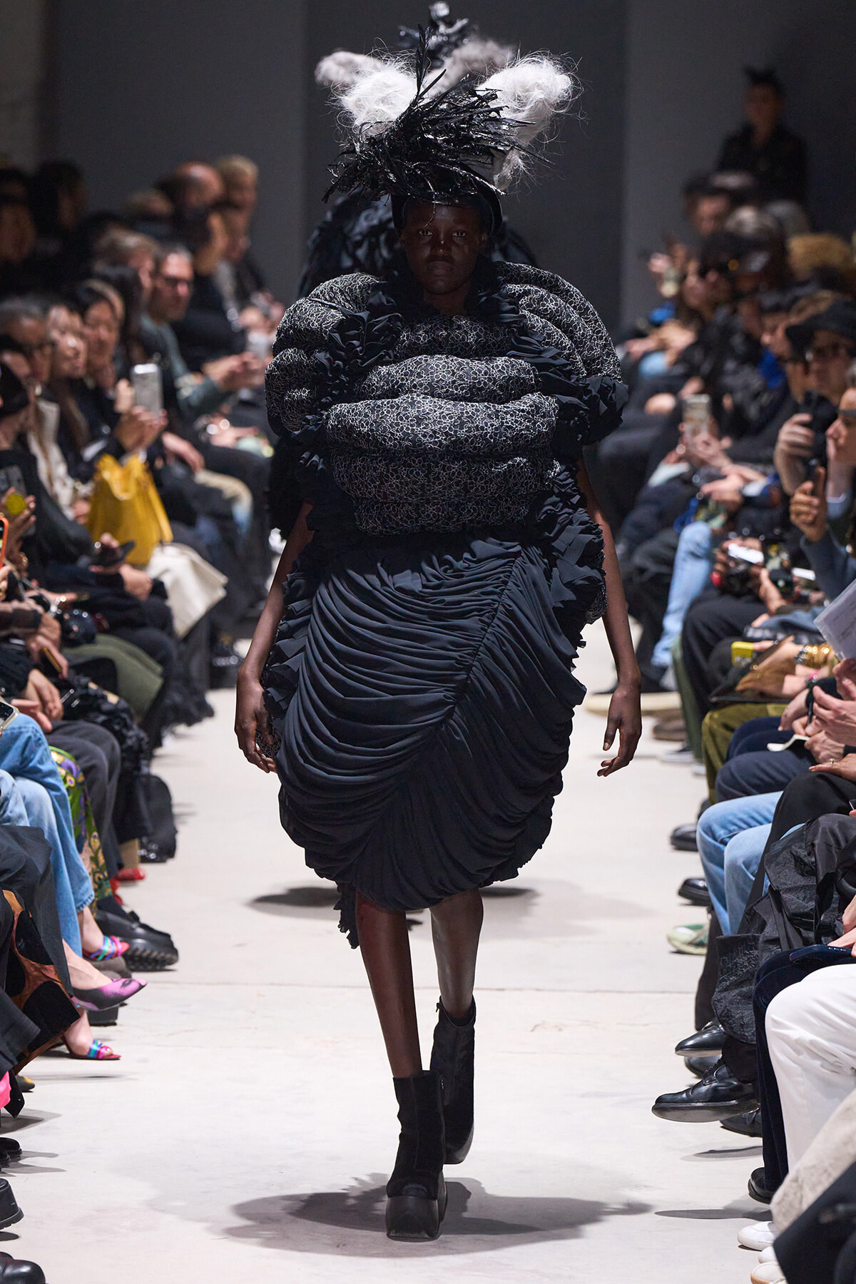 Model walks the runway in a dramatic black sculptural dress with a voluminous quilted bodice and feathered headpiece.