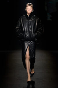 Runway model in an oversized black leather jacket and matching slit skirt, hands in pockets, on a dark catwalk.