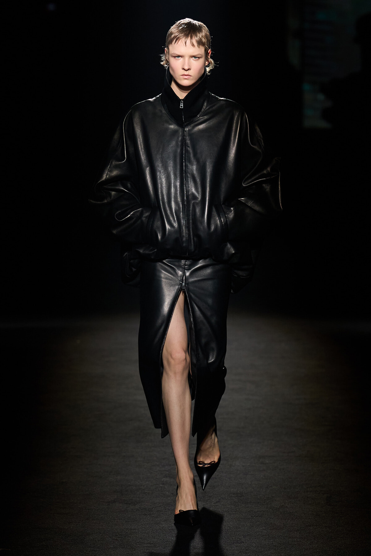 Runway model in an oversized black leather jacket and matching slit skirt, hands in pockets, on a dark catwalk.