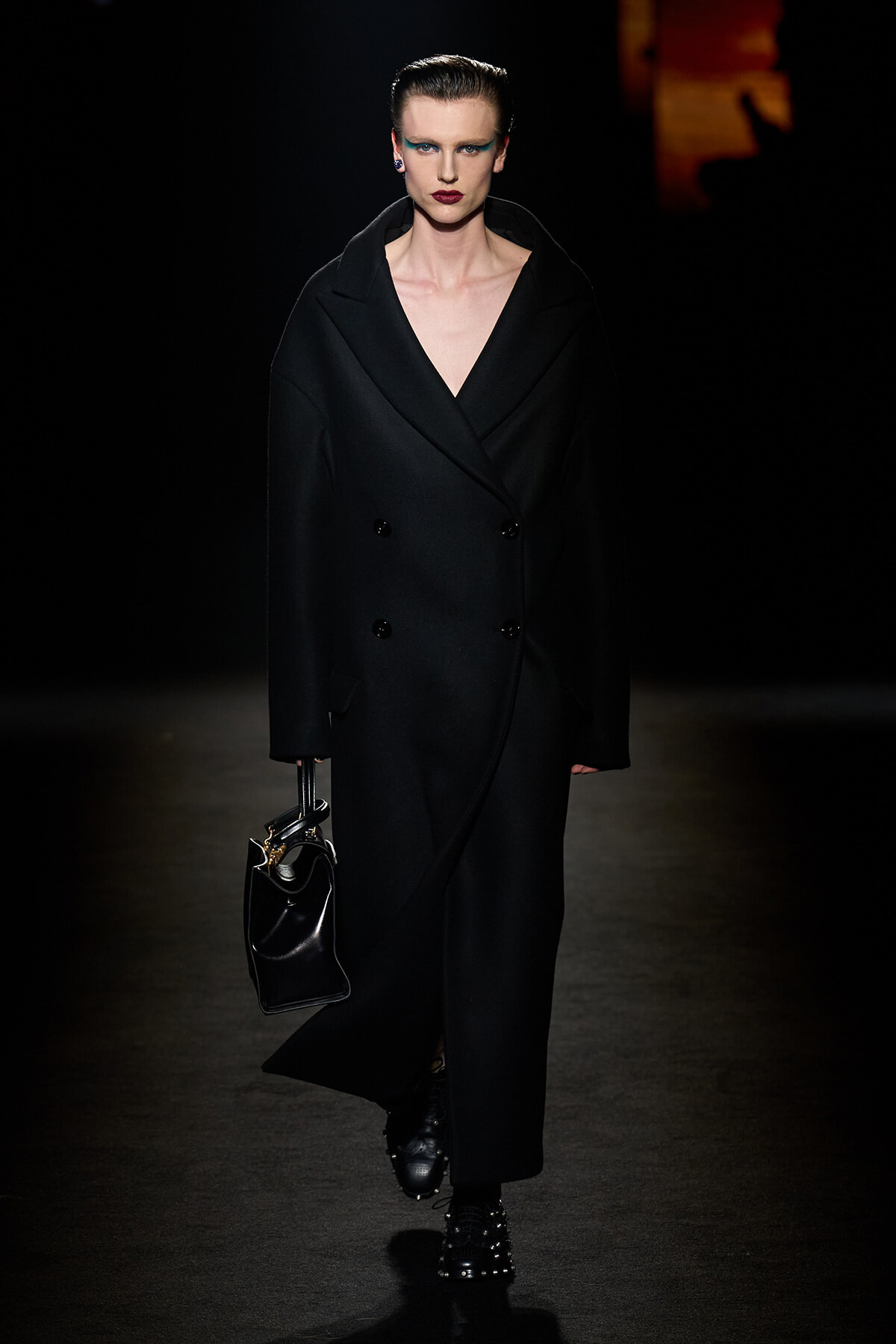 Model on a dark runway wearing an oversized black double-breasted coat, carrying a small black leather handbag.