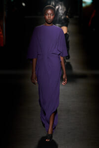 Model wearing a flowing purple dress on a dimly lit runway, hands at her sides.