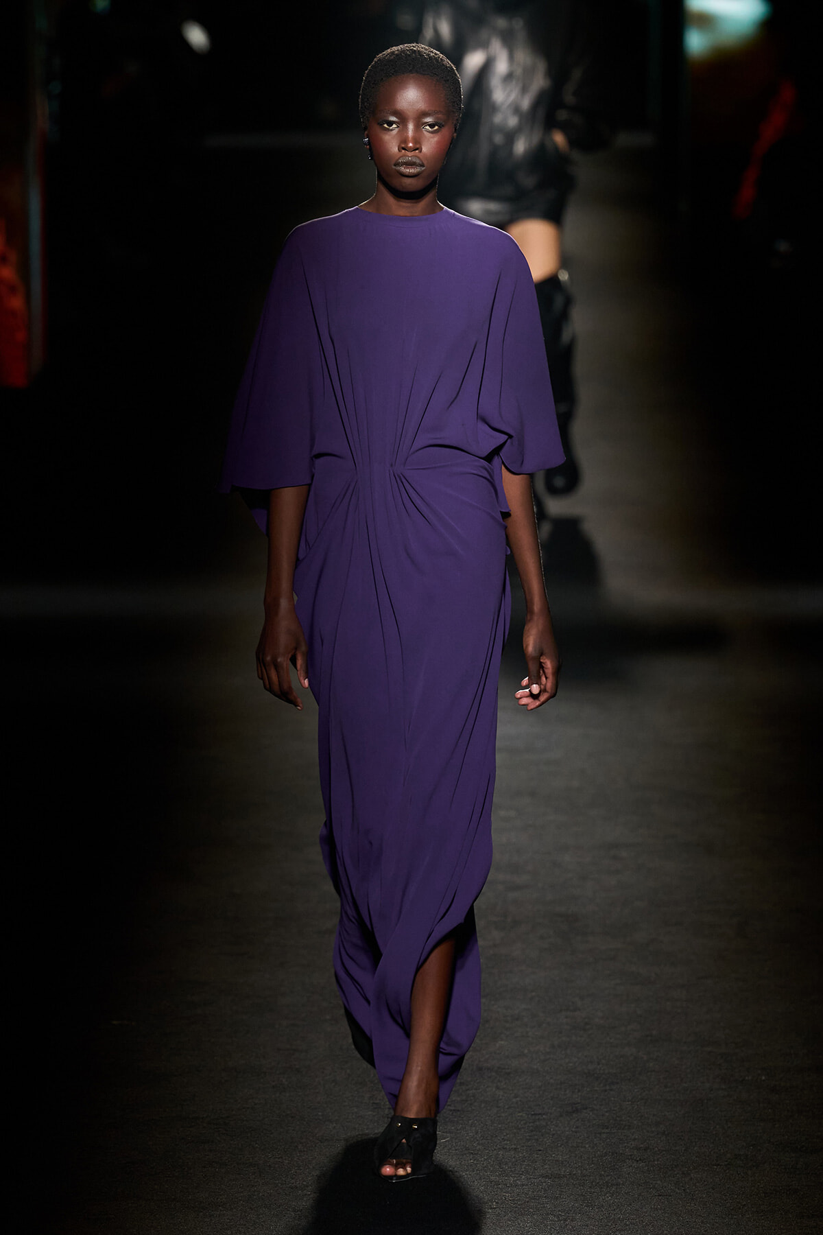 Model wearing a flowing purple dress on a dimly lit runway, hands at her sides.