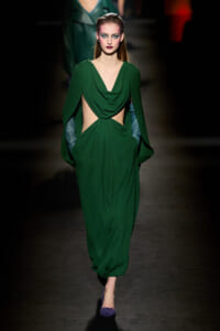 Female model walks the runway in a green draped gown with waist cutouts and long cape-like sleeves, purple shoes.