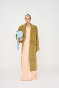 Older woman with gray hair wearing a long camel coat over a peach dress, holding a light blue handbag against a white studio background.