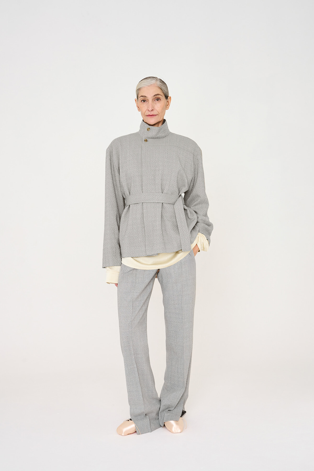 Full-body shot of a senior woman wearing a light gray belted knit suit with a high collar and cream inner layer, standing against a plain backdrop.