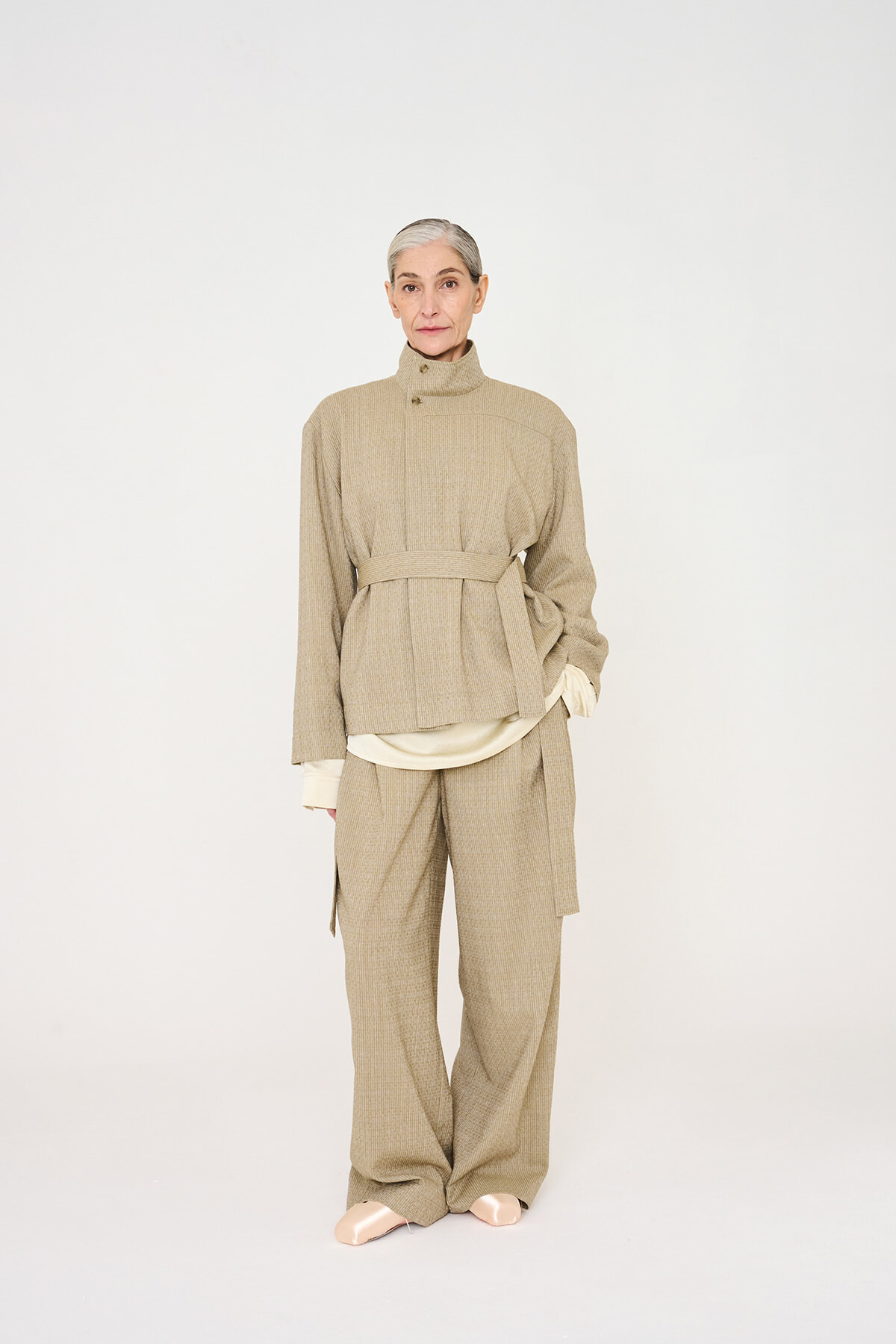 Model wearing a beige belted knit jacket and matching wide-leg pants in a neutral studio setting, tan-toned layered look.