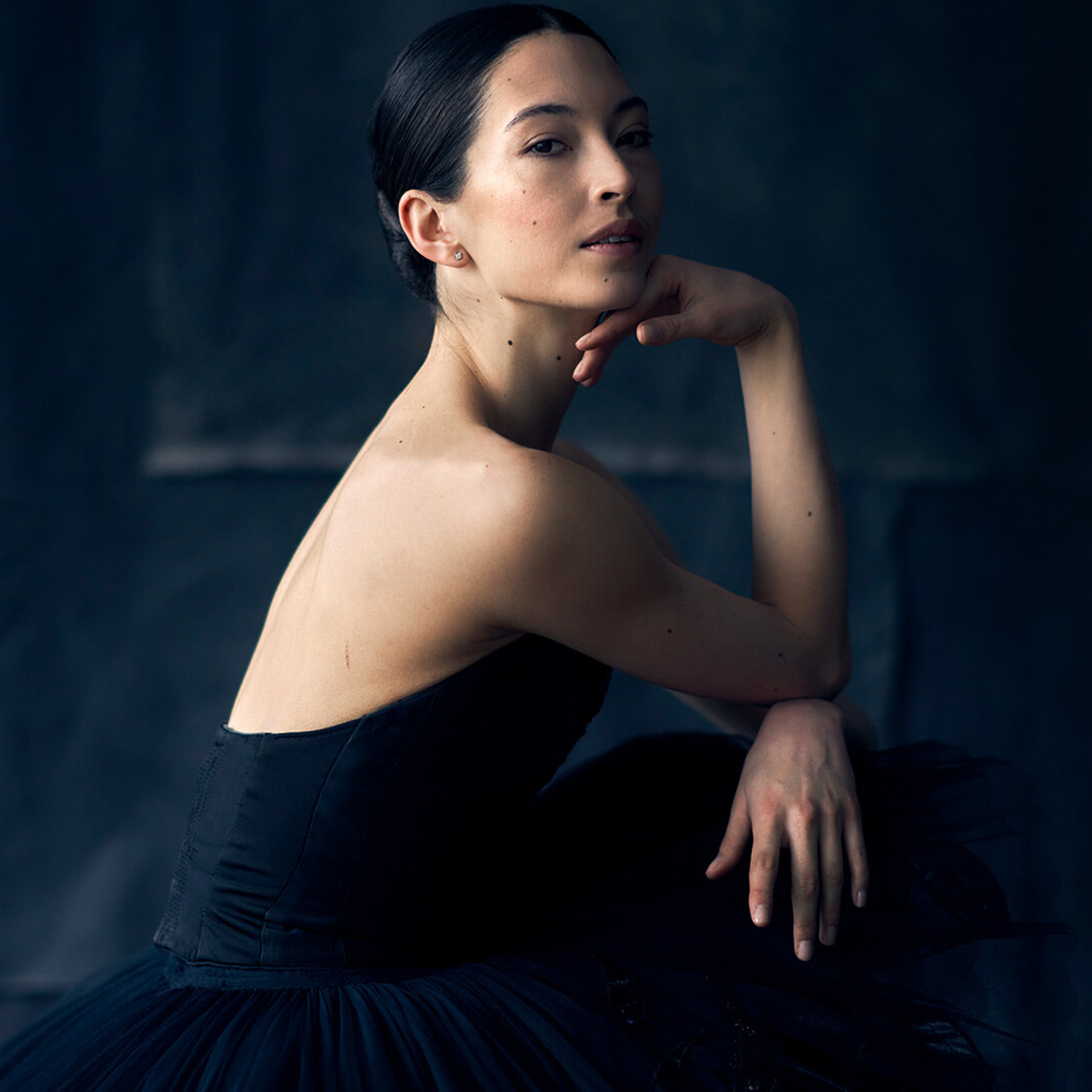 Portrait of a woman in a black strapless corset and tulle skirt, seated with her chin resting on her hand against a dark blue background.