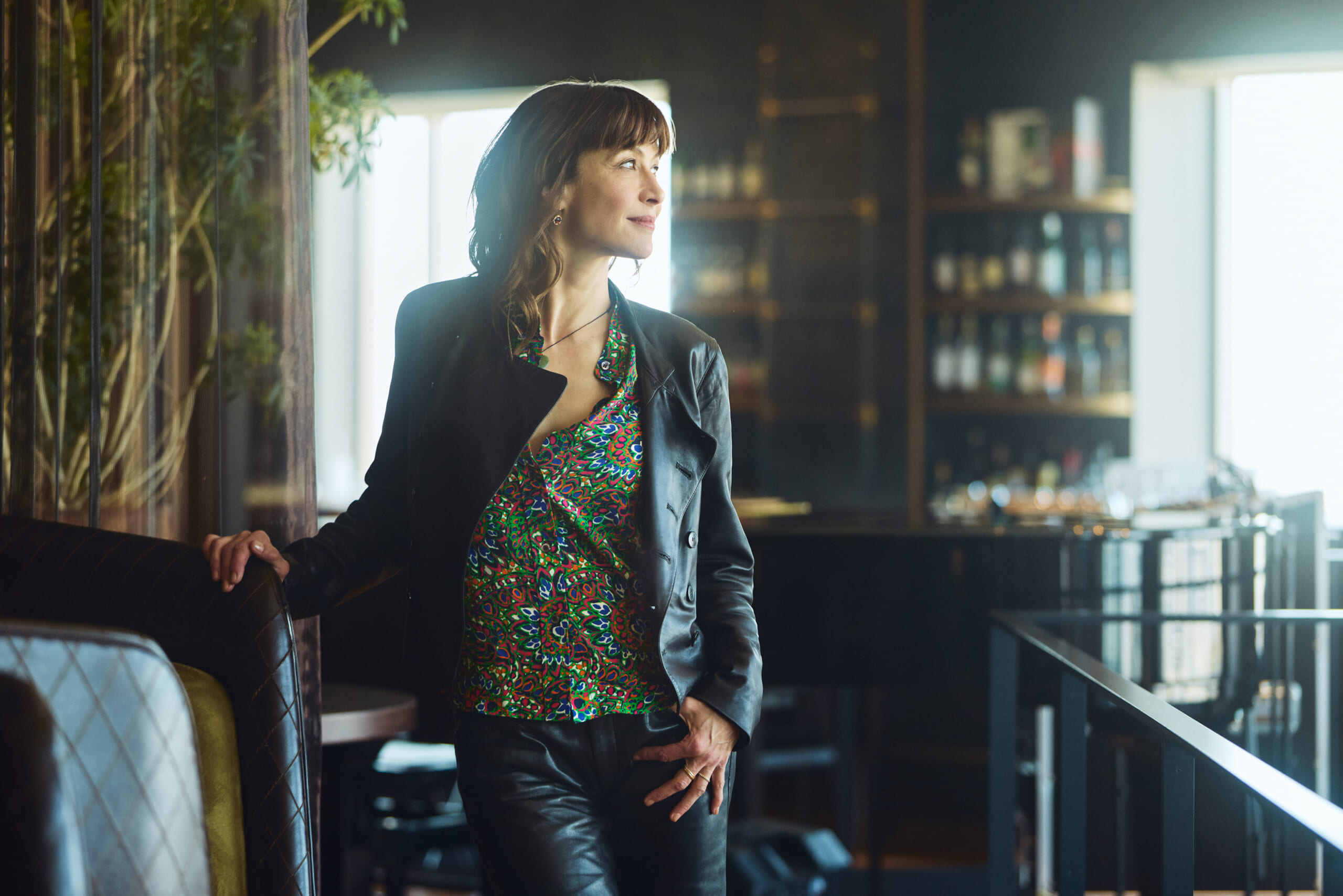 Woman in a colorful patterned blouse and black leather jacket stands leaning on a booth in a dim upscale bar, looking off to the side.