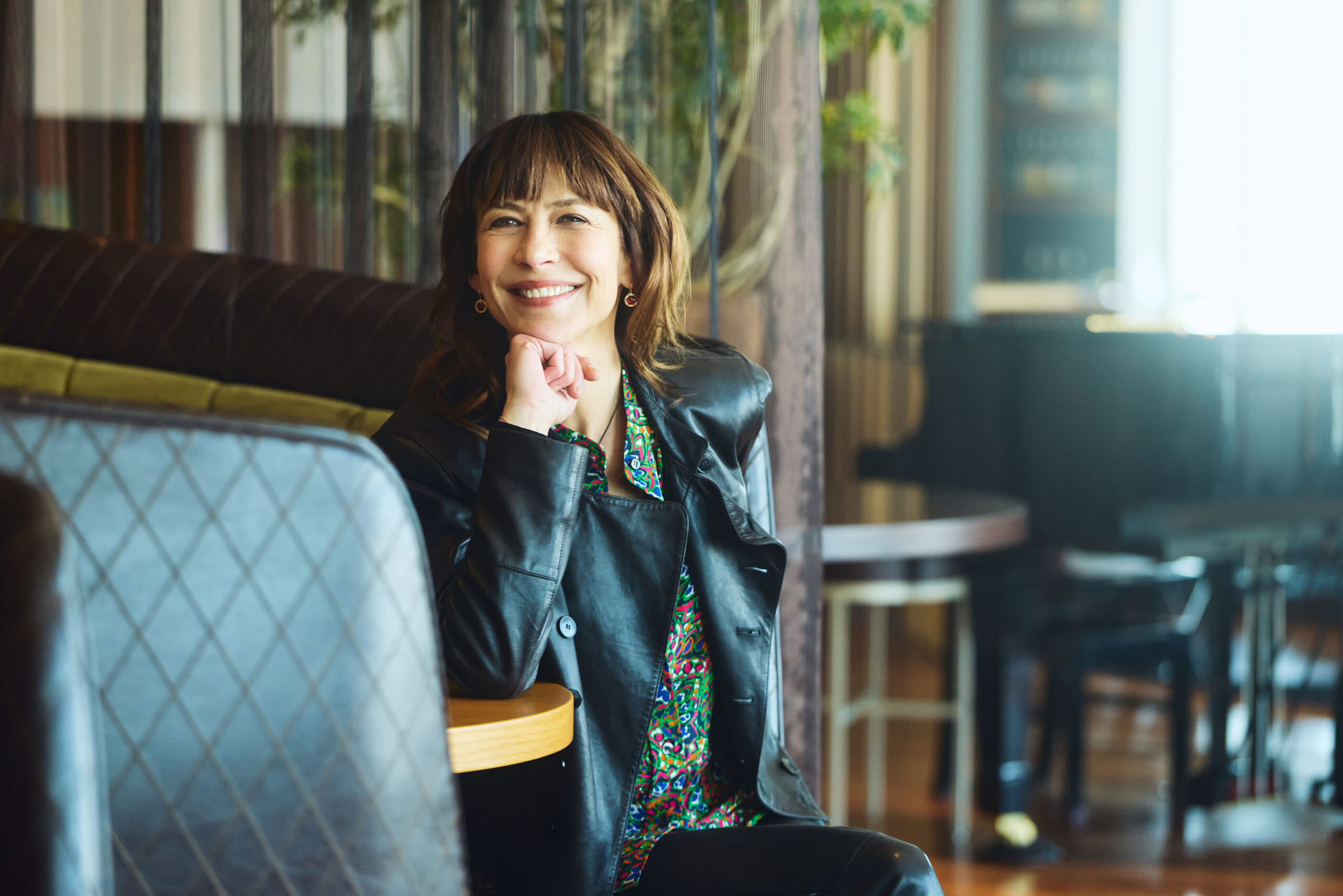 Smiling woman in a black leather jacket sits at a cafe table with a striped bench behind her.