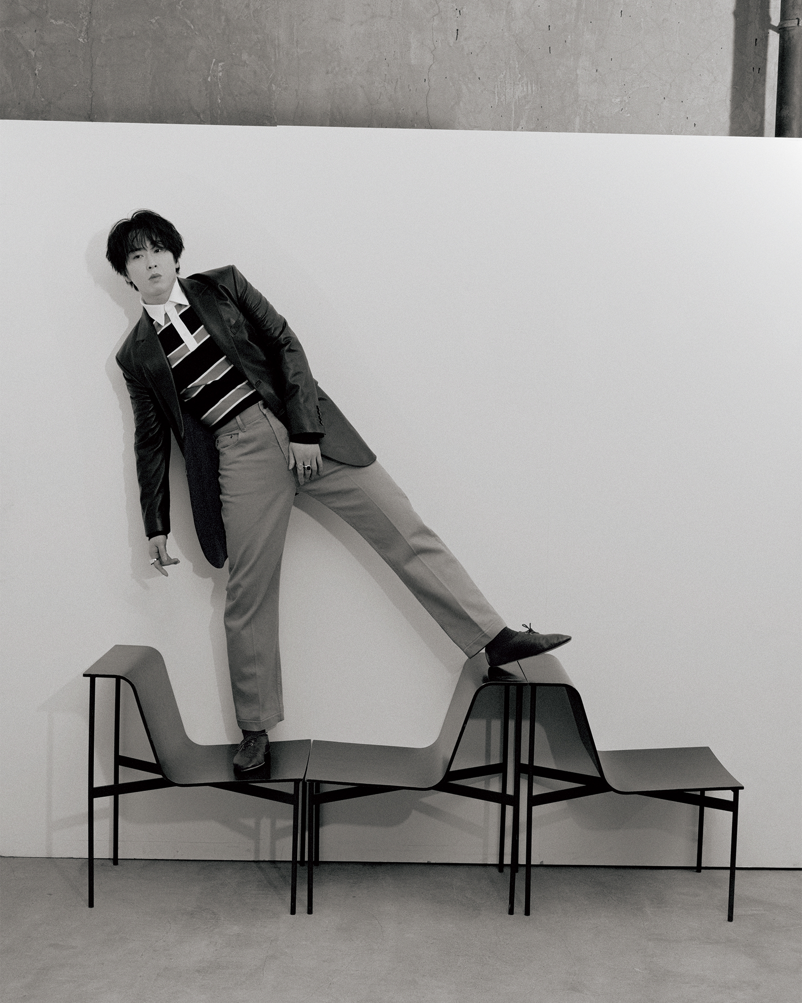Black-and-white fashion shot of a man in a blazer and striped sweater balancing with one foot on stacked chairs against a plain wall in a studio setting.