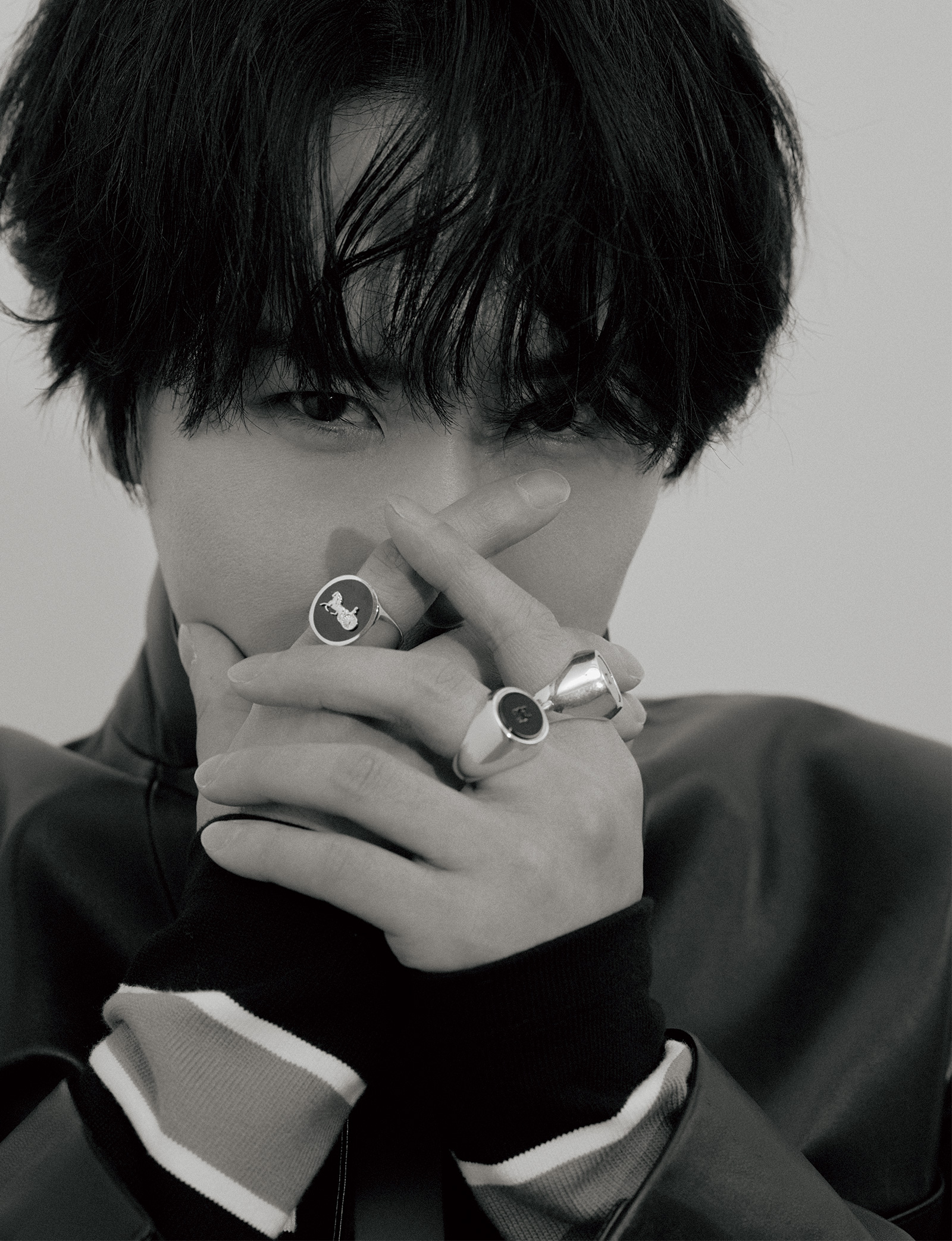 Close-up black-and-white portrait of a person with dark hair, hands crossed over their mouth, wearing bold rings on fingers.
