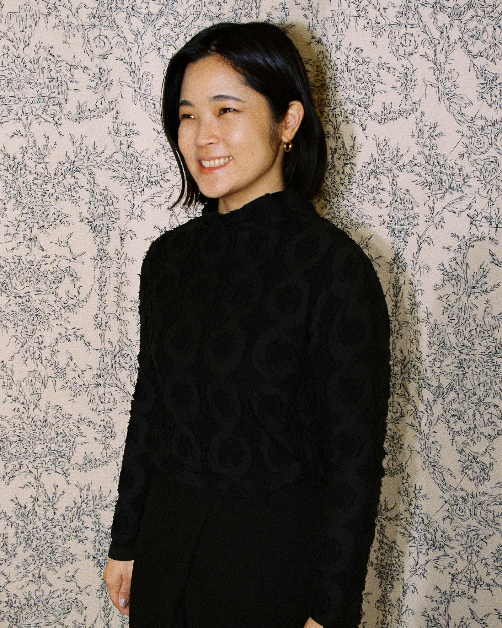 Portrait of a smiling woman with shoulder-length black hair in a black lace dress, standing against a pale floral-patterned wall.