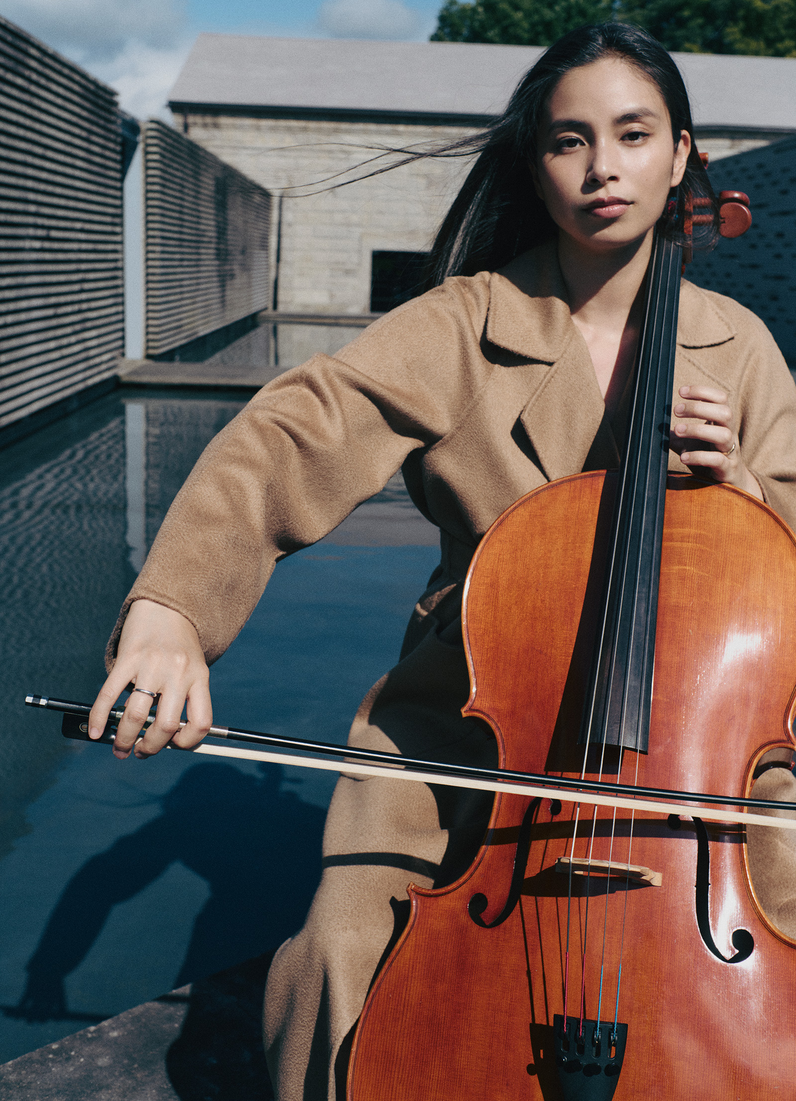 260831-yuiko-kawauchi-maxmara-01 | madameFIGARO.jp(フィガロジャポン) Woman playing a cello outdoors, bow in her left hand, wearing a tan coat.