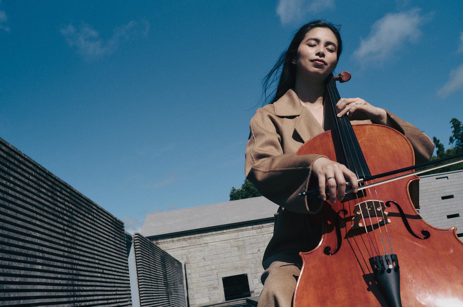260831-yuiko-kawauchi-maxmara-02 | madameFIGARO.jp(フィガロジャポン) Woman plays cello outdoors under a clear blue sky, wearing a tan coat while bowing the strings.
