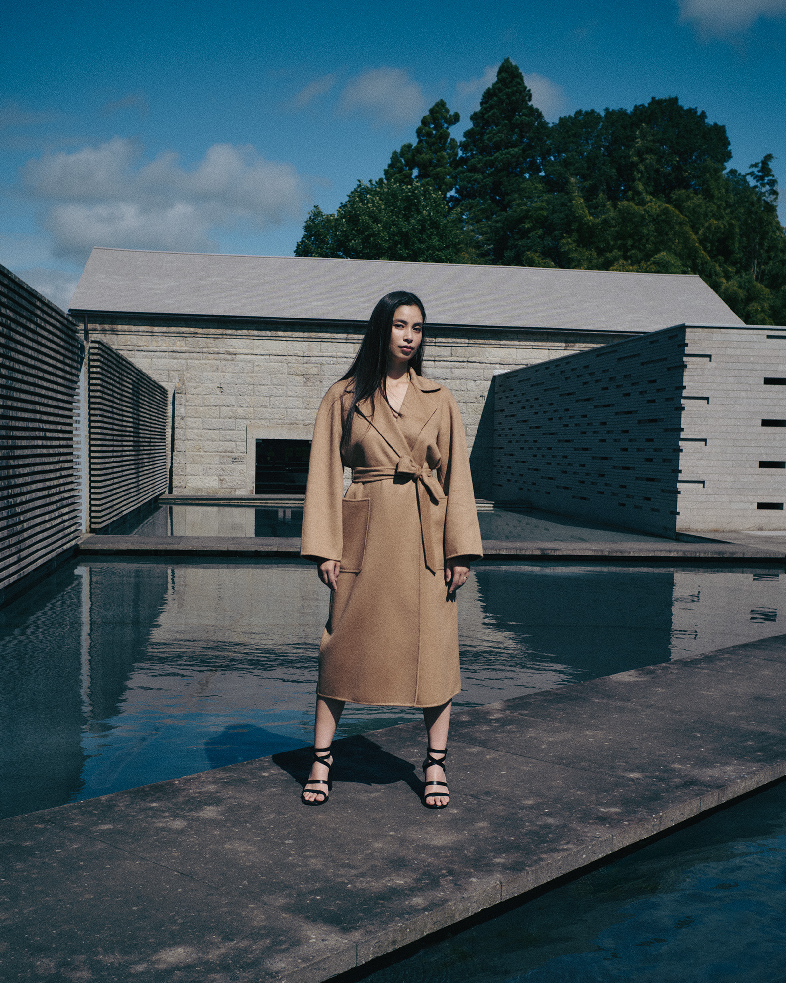 260831-yuiko-kawauchi-maxmara-03 | madameFIGARO.jp(フィガロジャポン) Woman in a tan coat standing by a reflective pool at a modern stone building.