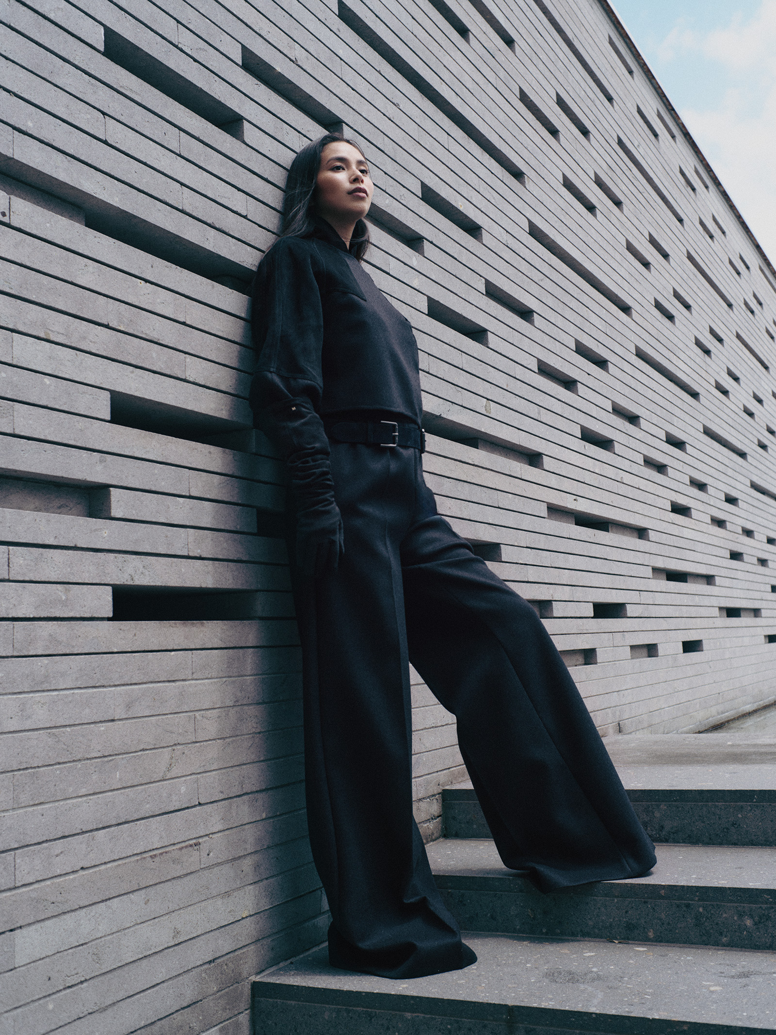 260831-yuiko-kawauchi-maxmara-04 | madameFIGARO.jp(フィガロジャポン) Woman in a black outfit leaning against a textured gray brick wall beside outdoor steps.