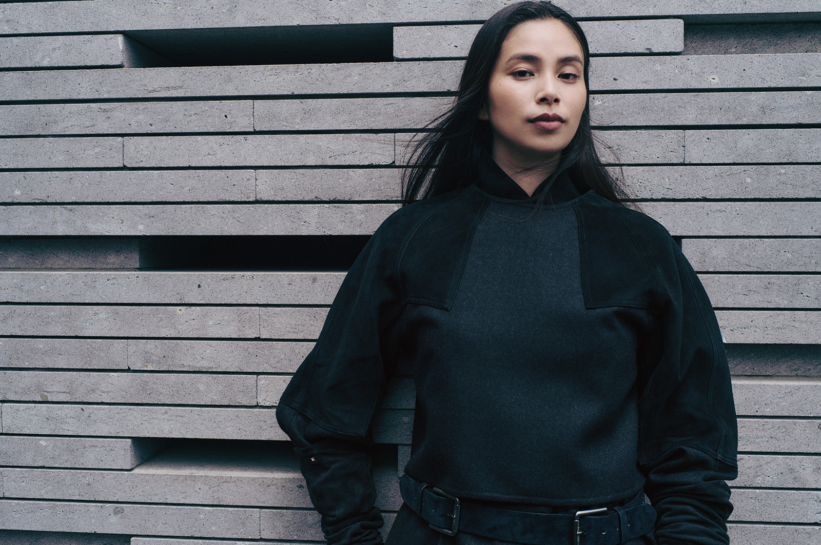 260831-yuiko-kawauchi-maxmara-05 | madameFIGARO.jp(フィガロジャポン) Woman with long dark hair in a black oversized sweater and belt, posing against a textured concrete wall with horizontal slats.