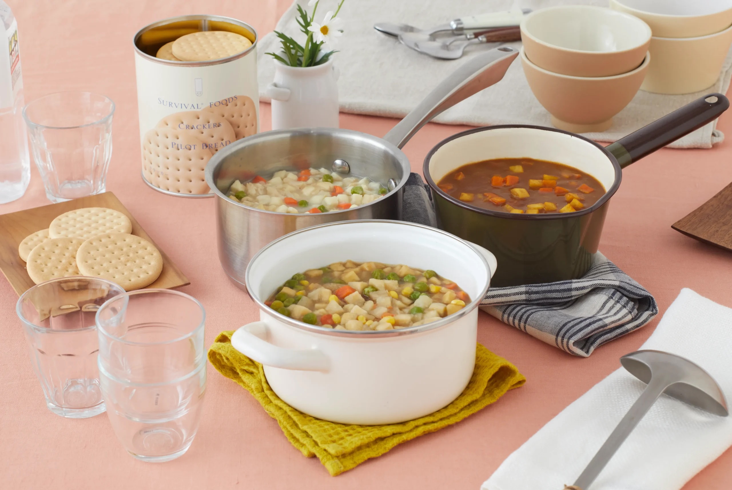 Table set for a meal: two saucepans of vegetables cooking in pots, a tin of crackers, stacked bowls, and clear glasses on a pink table.