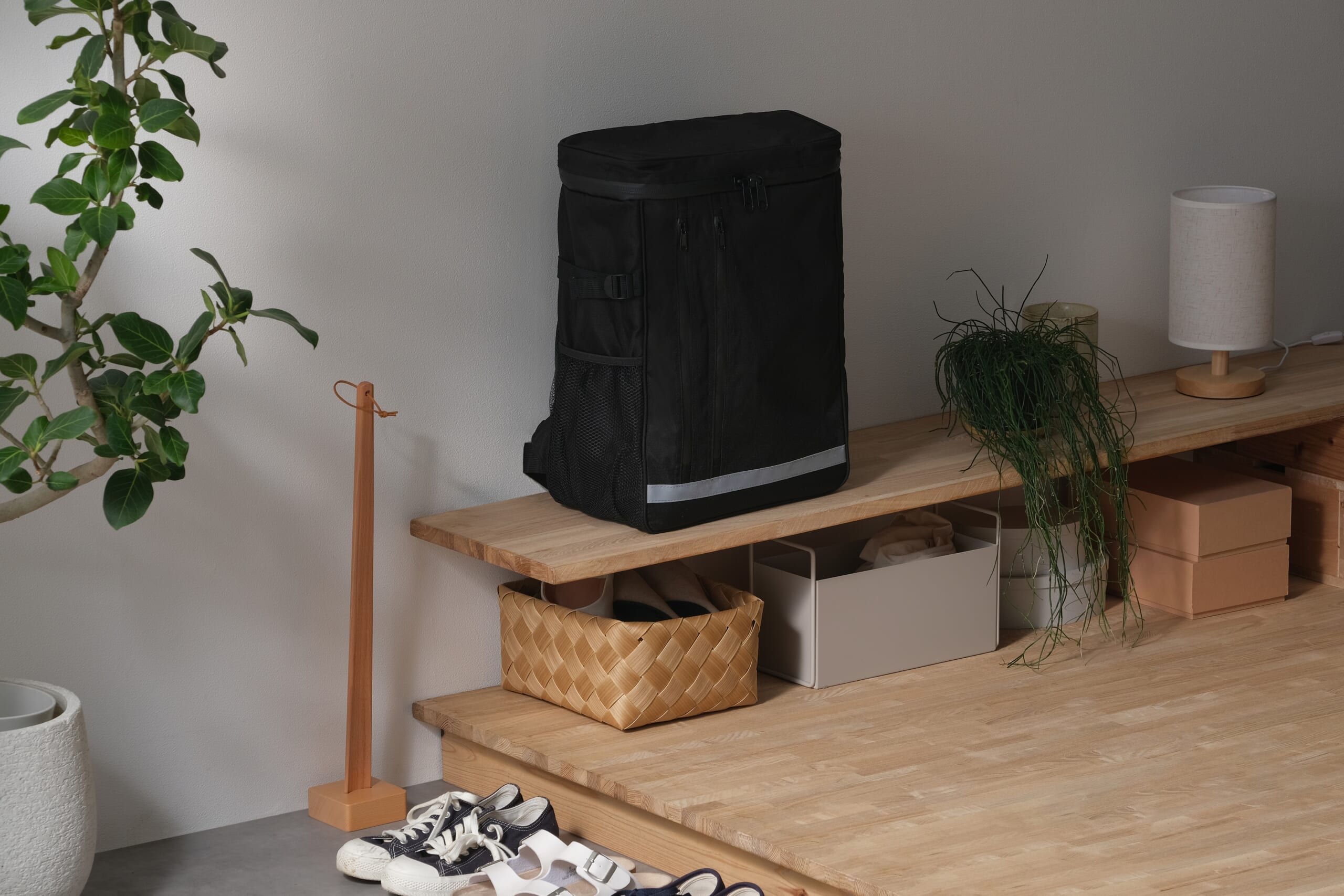 Black backpack resting on a wooden shelf with a green plant nearby and shoes on the floor in a tidy entryway or living area.