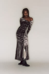 Dark-skinned woman wearing a metallic off-shoulder, ruched, floor-length dress with boots, posing against a light background.