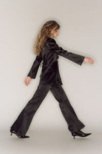 Person in a black satin suit (blazer and wide‑leg pants) walking forward against a neutral backdrop, showcasing motion and style.